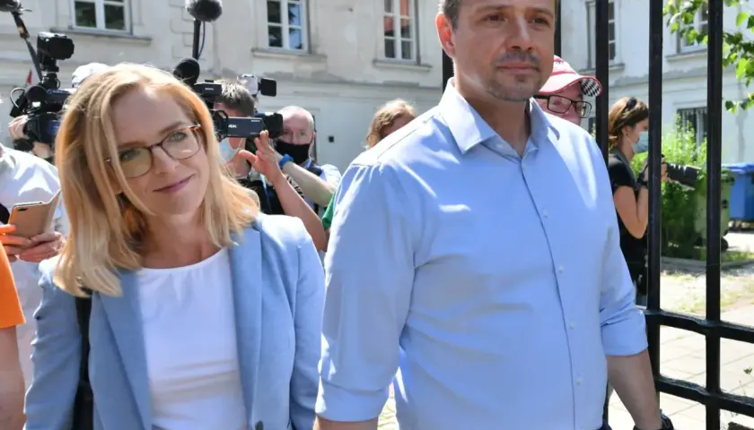 Małgorzata Trzaskowska w jasnej marynarce i Rafał Trzaskowski w błękitnej koszuli otoczeni przez dziennikarzy.