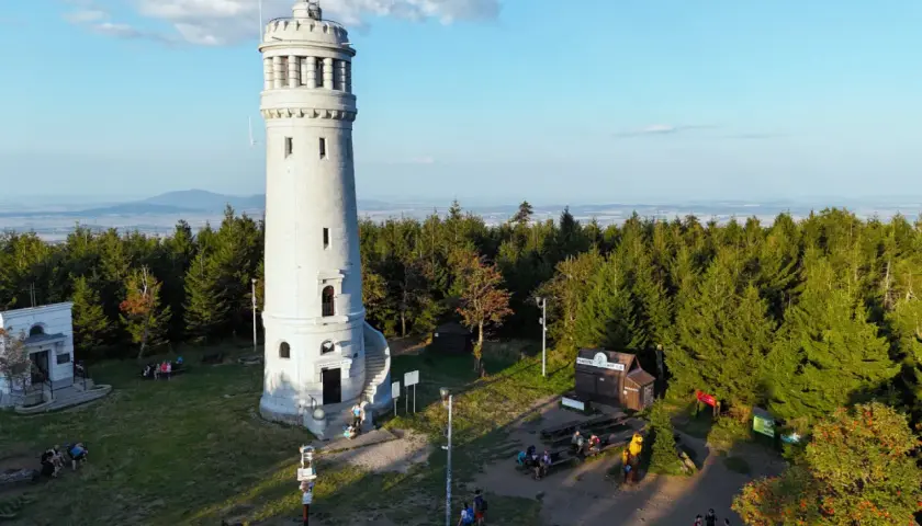 Wieża widokowa na szczycie góry, otoczona lasem. W dole mały domek i ławki, gdzie odpoczywają turyści. Wygląda jak wielka sowa czuwająca nad okolicą.