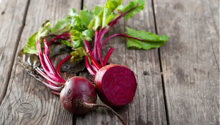 Zwei frische rote Bete, eine davon angeschnitten, liegen mit grünen Blättern auf Holz. Perfekt, um rote Beete roh zu essen.