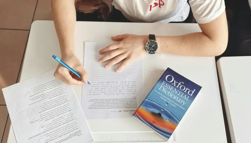 Studente che scrive, con un dizionario Oxford Essential Dictionary aperto. Cosa vuol dire AT? Forse "at" è un'abbreviazione usata in questo contesto di studio.