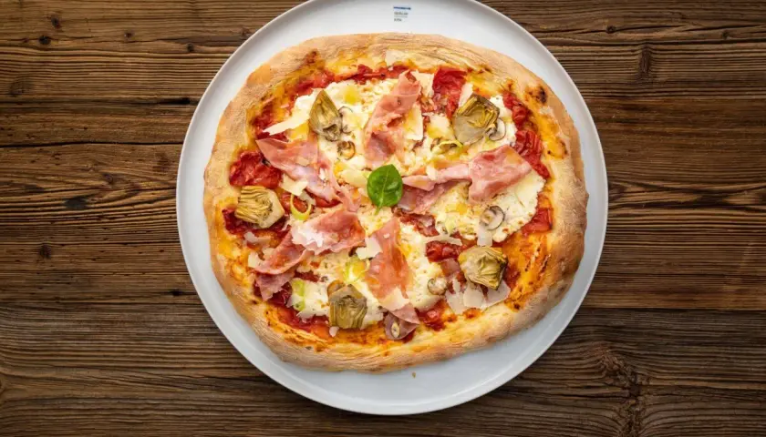 Eine köstliche Pizza mit Prosciutto Crudo, Artischocken und frischem Basilikum auf einem weißen Teller, serviert auf rustikalem Holz.