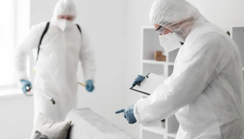 Dwóch specjalistów w kombinezonach ochronnych dezynfekuje mieszkanie. Jeden z nich trzyma opryskiwacz, drugi notuje na kartce. Może to być dezynfekcja po chorobie, lub użycie denaturatu do czyszczenia.