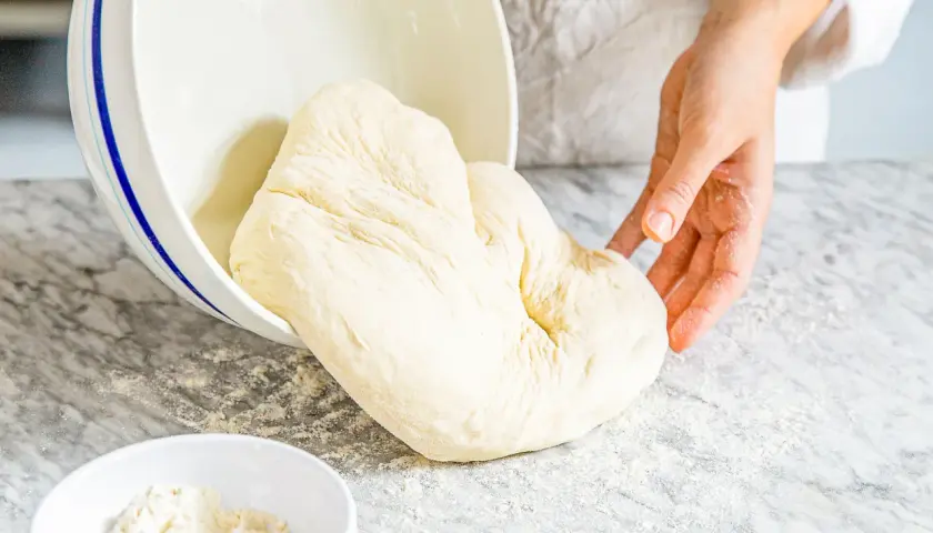 Frischer Pizzateig wird aus einer Schüssel auf eine bemehlte Arbeitsfläche gegeben. Das Mehl ist der Schlüssel für den perfekten Pizzaboden.