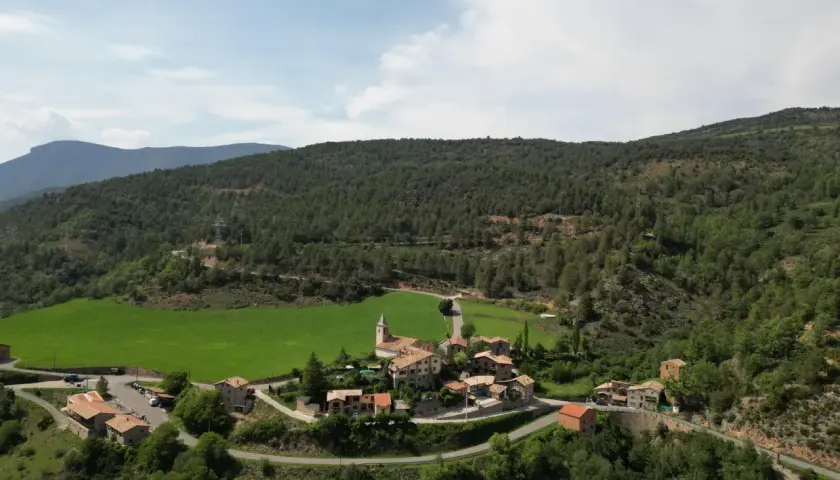 Vista aérea del pintoresco pueblo de **Vall de la Vansa**, rodeado de verdes prados y frondosos bosques, con casas de piedra y una iglesia.
