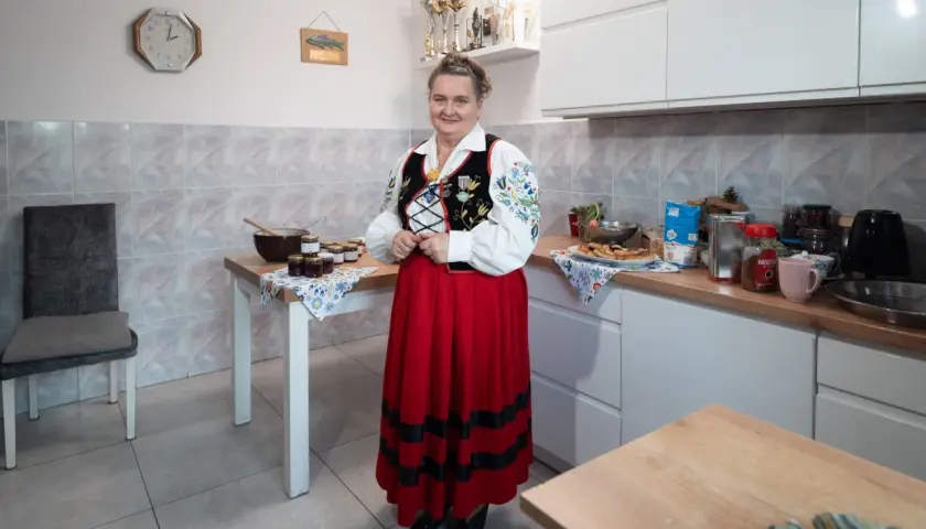 Kobieta w stroju ludowym stoi w kuchni obok stołu z dżemami. Na ścianie wisi zegar, wskazujący godzinę 10.
