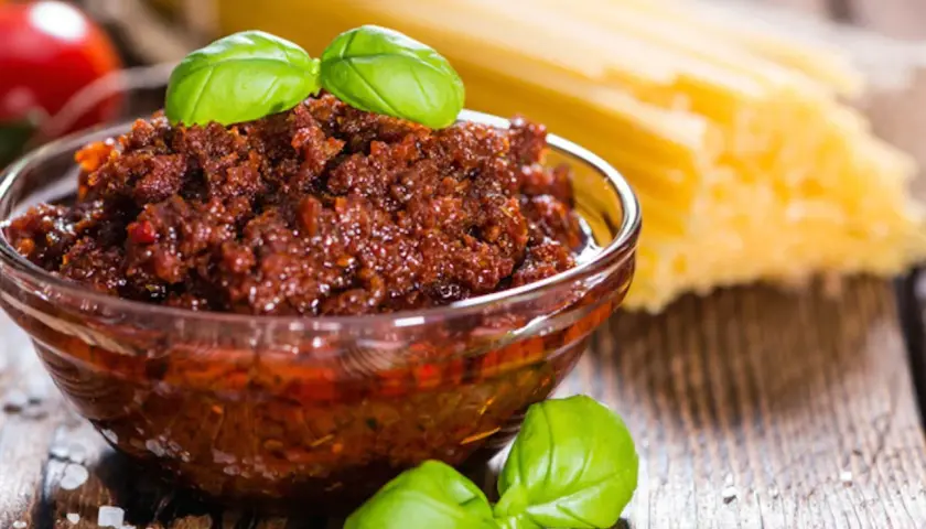 Pesto di pomodori secchi in una ciotola di vetro, con spaghetti e basilico sullo sfondo.