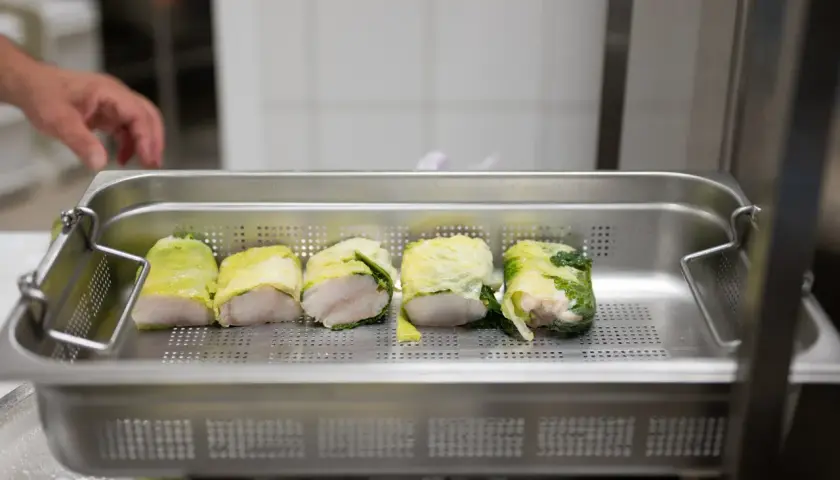 Cinq rouleaux de poisson enroulés dans des feuilles de chou, prêts pour la cuisson. Une main est visible en arrière-plan, préparant ce plat pour la journée internationale des cuisiniers.