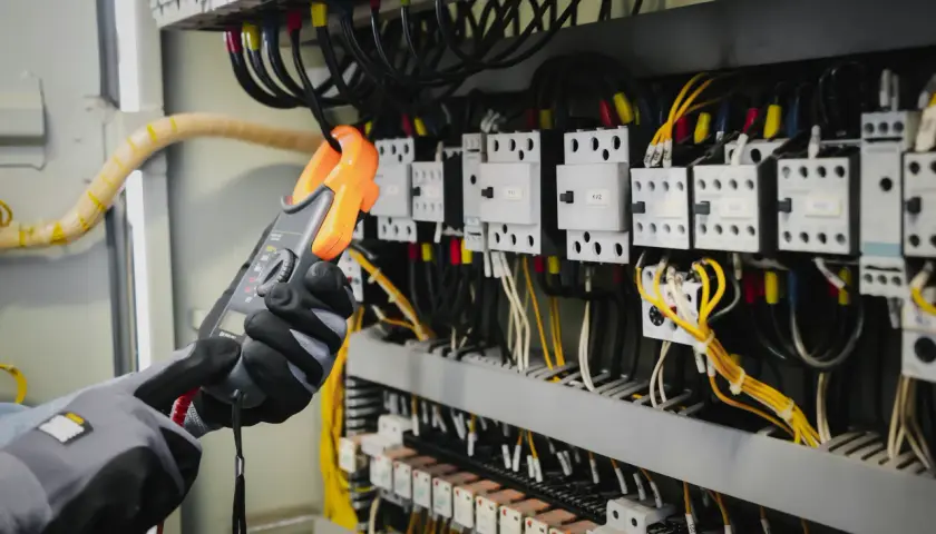 Ręce w rękawicach trzymają pomarańczowy miernik, badając skomplikowany opis rozdzielni elektrycznej z licznymi przewodami i przekaźnikami.