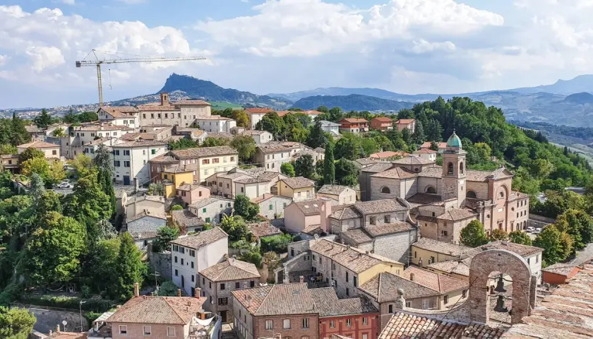 Panoramica di un borgo collinare con case dai tetti rossi, una chiesa con campanile e montagne sullo sfondo. Un paesaggio incantevole nei dintorni di Rimini.