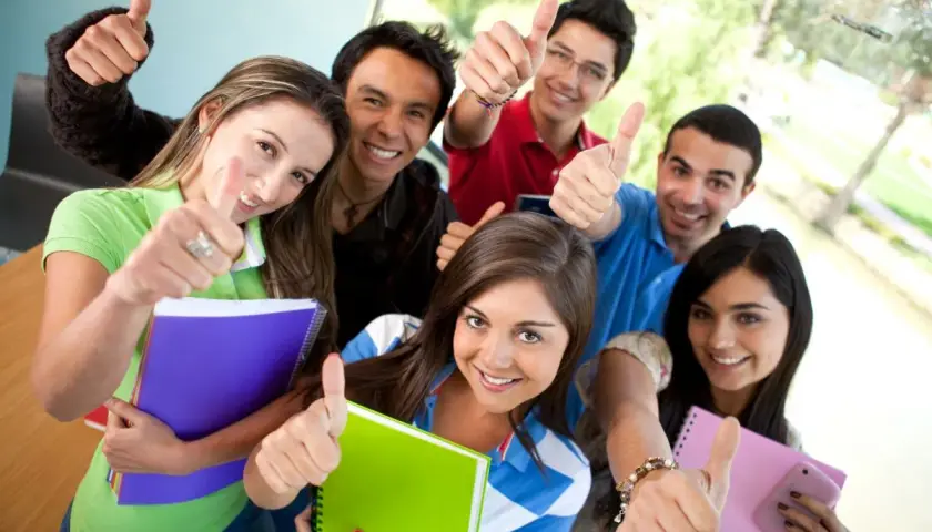 Uśmiechnięci studenci z notatnikami pokazują kciuki w górę, gotowi na studia stacjonarne.