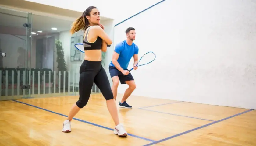 Para ćwiczy squasha, poznając zasady gry. Kobieta w sportowym stroju i mężczyzna w niebieskiej koszulce z rakietami gotowi do akcji na boisku.