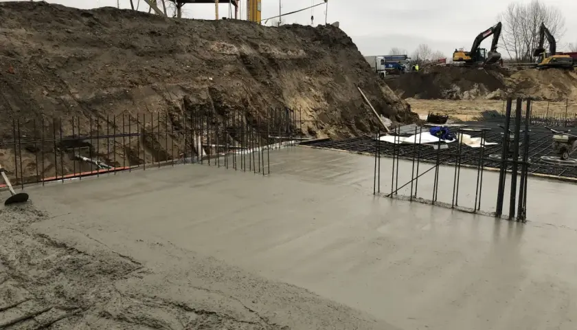 Budowa w toku: świeża wylewka betonowa na zewnątrz, zbrojenie i koparki w tle.