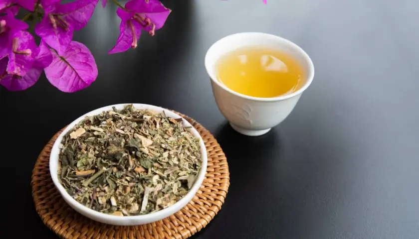 Una tazza di tisana per digerire accanto a una ciotola di erbe secche, con fiori viola sullo sfondo.