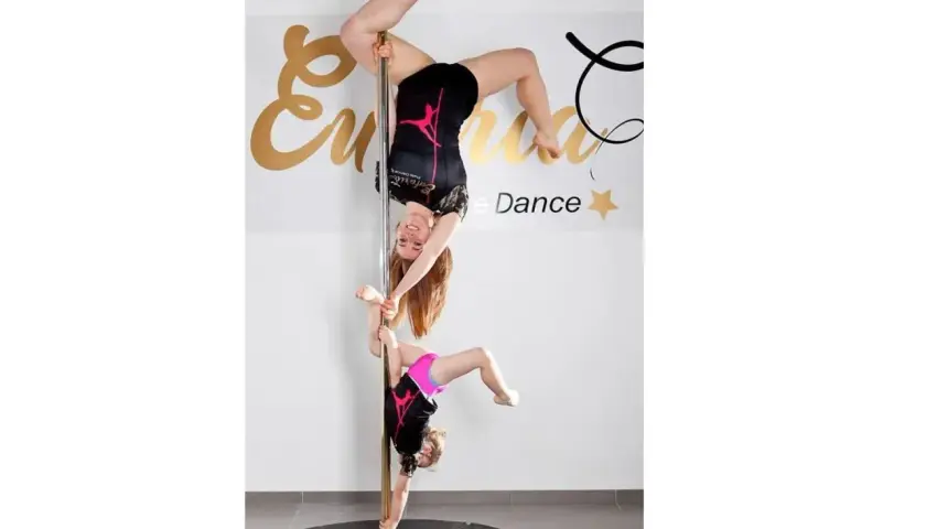 Dwie dziewczyny ćwiczą pole dance. Młodsza wspiera starszą, która wisi głową w dół.