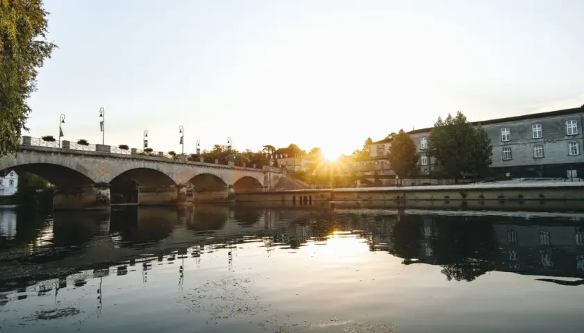 Un pont de pierre traverse une rivière calme, reflétant le soleil couchant sur les bâtiments historiques. Une belle vue pour savoir quoi faire à Cognac.