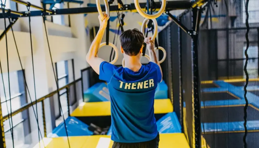 Trener w niebieskiej koszulce z napisem "TRENER" trzyma się kółek gimnastycznych. W tle widać trampoliny i siatki. To miejsce, gdzie można wzmocnić powięź.