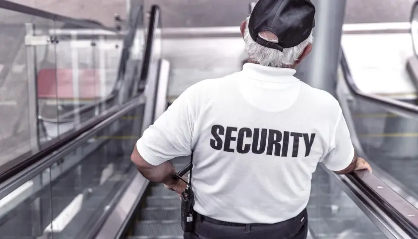 Doświadczony technik ochrony fizycznej osób i mienia, w białej koszulce z napisem "SECURITY", stoi na ruchomych schodach.
