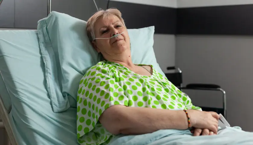 Starsza kobieta w szpitalnym łóżku, z tlenem przy nosie, w zielono-białej bluzce w groszki. Czeka na leki, być może Duracef.