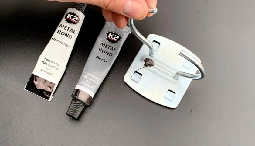 Dwie tubki kleju do blachy K2 Metal Bond (żywica i utwardzacz) oraz metalowy element z nałożonym klejem.