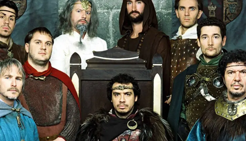 Le casting de Kaamelott réuni, des chevaliers aux allures diverses, certains sur un trône, d'autres en armure.