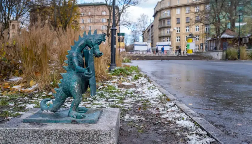 Mały smok z brązu, symbol smoczy szlak kraków, stoi na kamiennym postumencie. W tle miejski krajobraz z budynkami i mokrą nawierzchnią.
