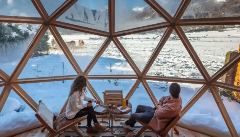 Pareja disfruta de un paisaje nevado en el Pirineo francés desde una cúpula geodésica. Vacas pastan en la distancia.