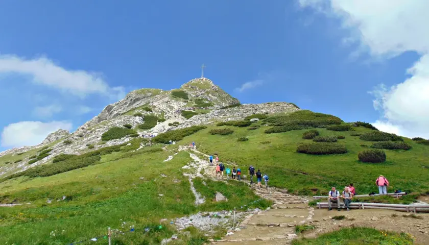 Tłum ludzi wspina się po kamienistym szlaku na Giewont. Widać krzyż na szczycie.