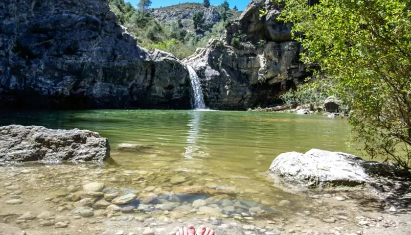Pies descalzos en la orilla del **barranco de la Encantada**, con una cascada cayendo en una poza de agua verde rodeada de rocas y vegetación.