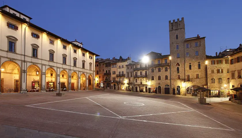 Piazza illuminata ad Arezzo, con portici, edifici storici e una torre. Sembra proprio che la vita è bella ad Arezzo.