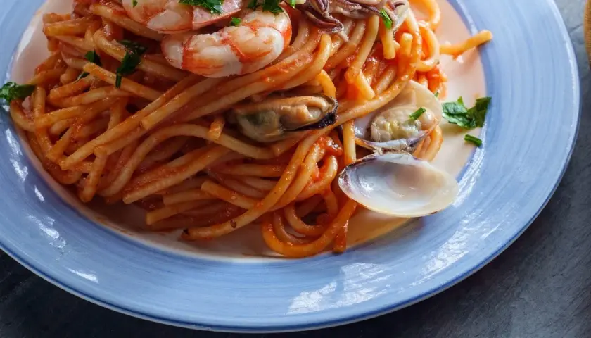 Espaguetis a la marinera con gambas, mejillones y almejas en una salsa roja, servidos en un plato azul.