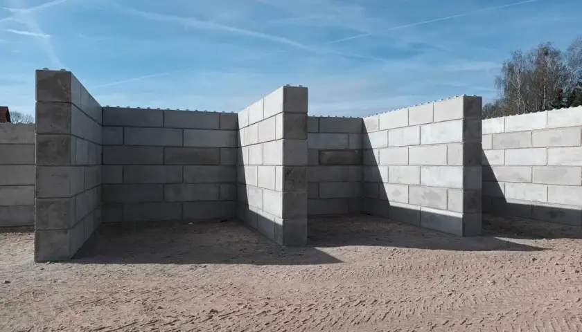 Budowa muru oporowego z bloczków betonowych. Konstrukcja tworzy zarys pomieszczeń na piaszczystym placu budowy.