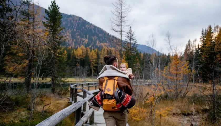 Ojciec z dzieckiem na plecach spaceruje drewnianą kładką pośród jesiennych lasów. Tatry z dzieckiem to piękna przygoda.