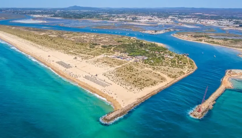 Vista aérea de la **isla de Tavira**, con su extensa playa de arena dorada, aguas turquesas y el canal que la separa de tierra firme, donde se aprecian salinas y la ciudad al fondo.