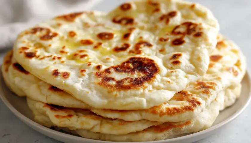 En hög med nybakade, gyllene naanbröd, perfekt för ditt nästa naan recept. Smält smör glänser på de fluffiga bröden.