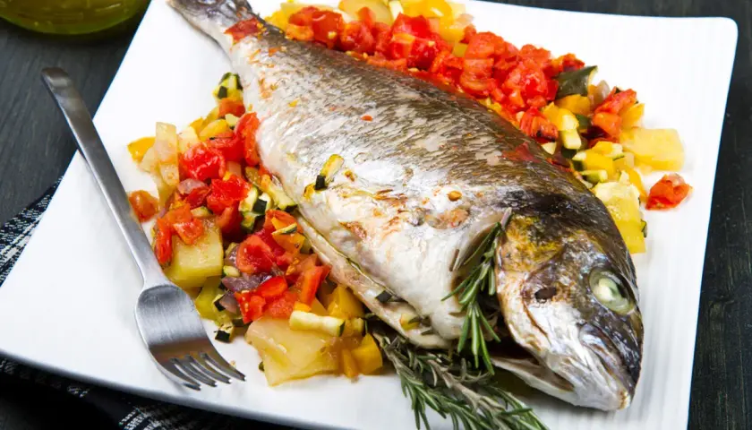 Dorada al horno con verduras asadas, servida en plato blanco con tenedor y romero fresco.