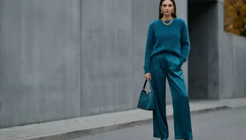 Elegancka kobieta w stylizacji w kolorze petrol, z błyszczącymi spodniami i miękkim swetrem, pozuje na tle betonowej ściany.