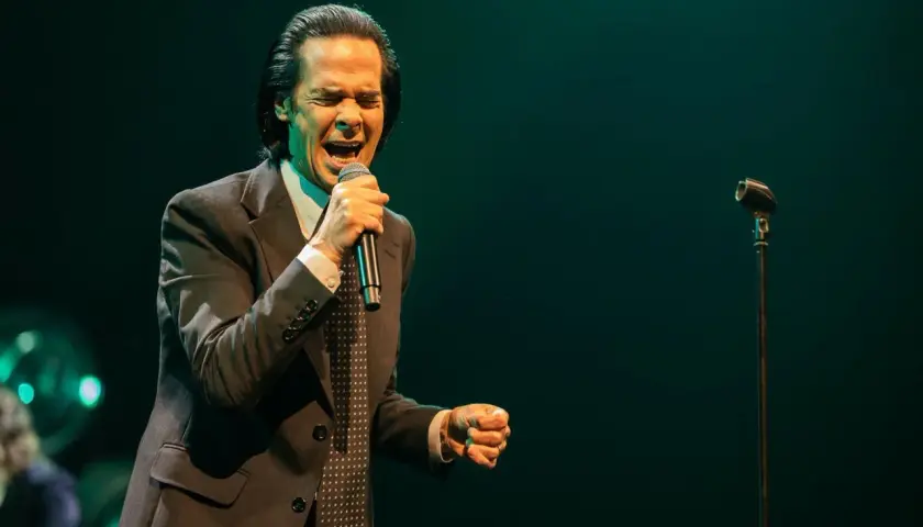 Nick Cave z pasją wykonuje swoje utwory, śpiewając do mikrofonu w eleganckim garniturze.