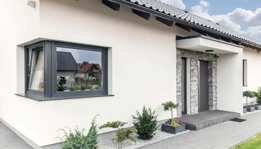 Nowoczesny dom z białym tynkiem, szarym dachem i efektownym **oknem narożnym ze słupkiem**. Przed wejściem ozdobne krzewy i kamyczki.