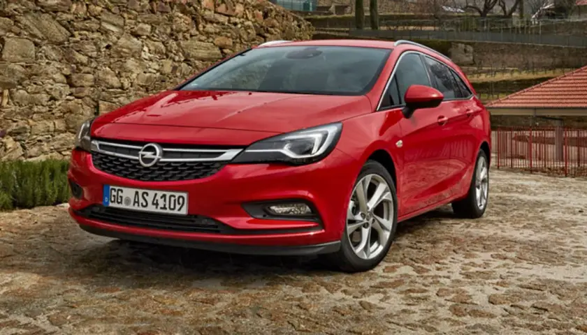 Czerwony Opel Astra Sports Tourer zaparkowany na kamienistej nawierzchni, z kamiennym murem w tle.