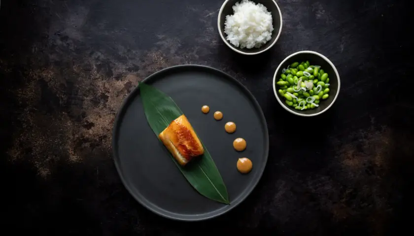 A perfectly cooked nobu miso black cod rests on a dark plate with a green leaf. Drizzled sauce and sides of rice and edamame complete the elegant dish.