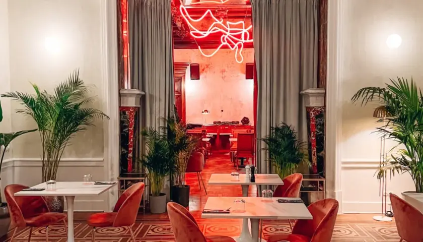 Eleganckie wnętrze z czerwonymi aksamitnymi krzesłami i marmurowymi stolikami. Neonowy napis w głębi sugeruje, że to najdroższa restauracja w Warszawie.