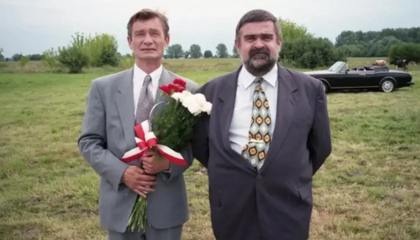 Wspólnik kilera z komedii Machulskiego, z bukietem kwiatów, obok kolegi i czarnego kabrioletu na łące.