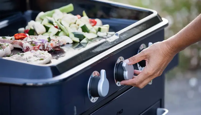 Main d'une personne tournant le bouton pour allumer plancha gaz. Viande et légumes coupés sont prêts à cuire sur la plaque.