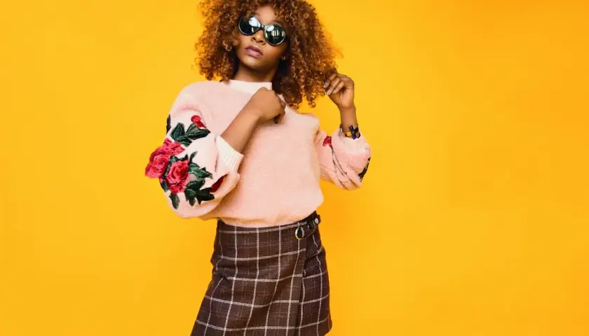 Mujer con cabello rizado y gafas de sol posa con un suéter rosa con rosas bordadas y una falda a cuadros. Su estilo es un armario minimalista moderno.