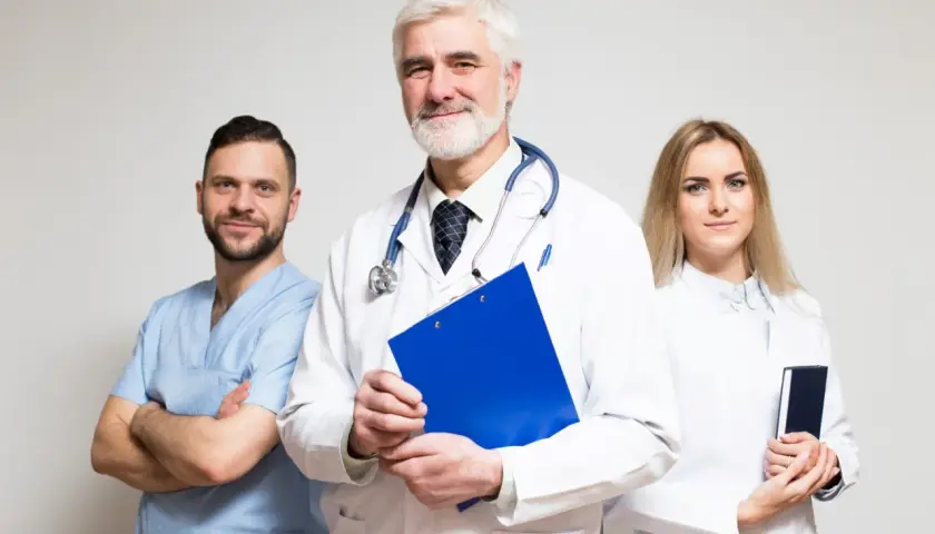 Tre medici sorridono, uno con camice azzurro, uno con camice bianco e stetoscopio, e una donna con camice bianco. Gestiscono la fatturazione medici con professionalità.