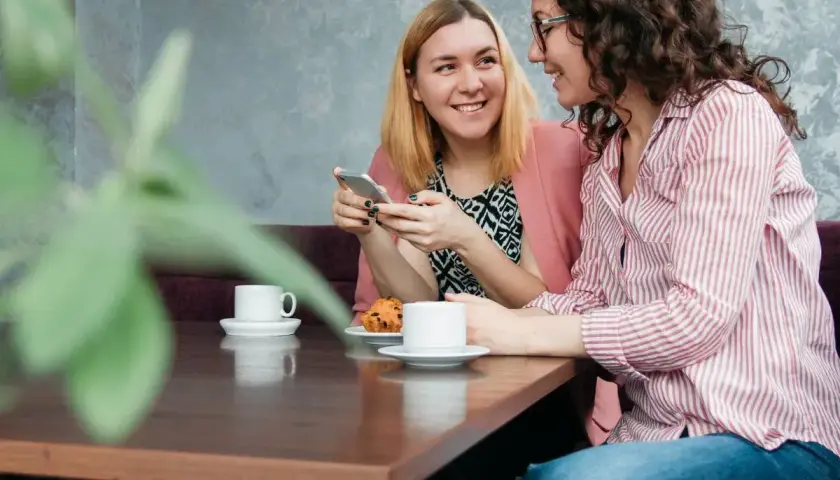 Dwie kobiety rozmawiają przy kawie, tworząc intymne tête-à-tête. Jedna pokazuje coś na telefonie, druga uśmiecha się.