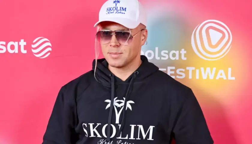 Skolim, Król Latino, w czarnej bluzie z kapturem i białej czapce, emanuje barwami szczęścia na tle festiwalowej ścianki.