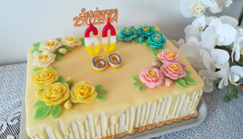 Żółty tort z ozdobami kwiatowymi i świeczkami w kształcie cyfr 60.