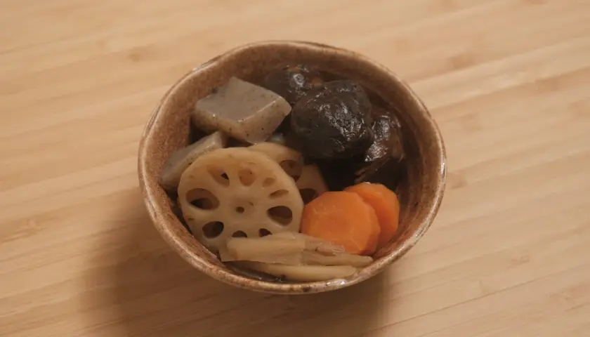 A bowl of simmered vegetables, including shiitake mushrooms, lotus root, carrots, and konjac, likely part of a mushroom dashi broth.