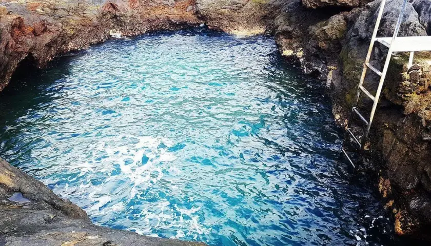Un charco de los abrigos natural, con aguas cristalinas y una escalera metálica para acceder. Rodeado de rocas volcánicas.